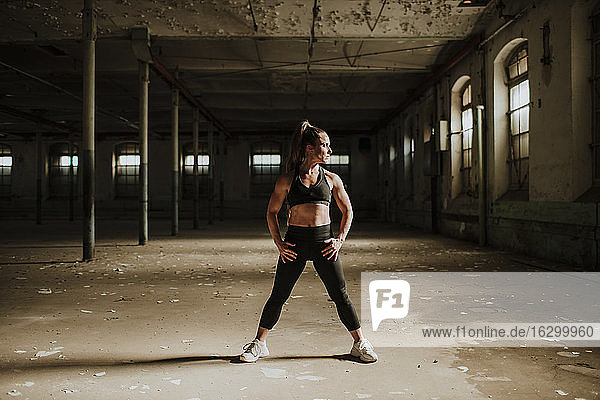 Muskulös gebaute Frau steht mit der Hand auf der Hüfte in einer verlassenen Fabrik