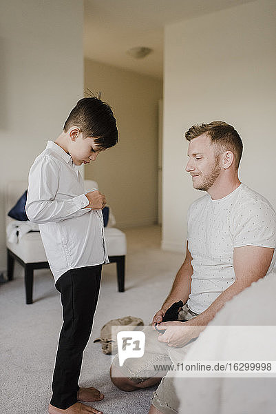 Smiling father kneeling by son getting dressed for wedding at home