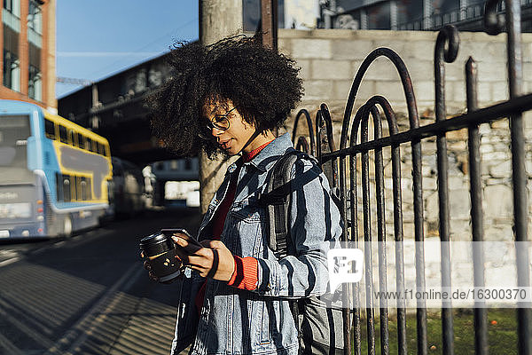 Young woman with curly hair using smart phone while standing by railing in city