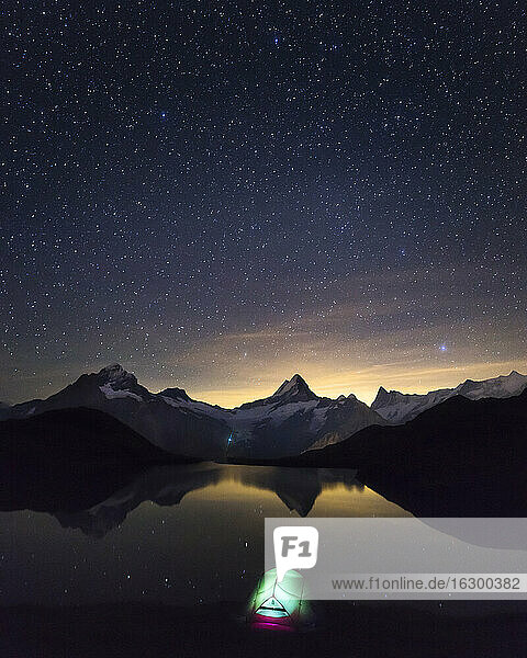 Starry night sky over illuminated tent pitched on shore of Bachalpsee lake at night