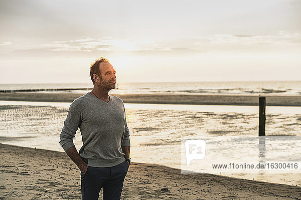 Thoughtful man with hands in pockets standing against sea during sunset