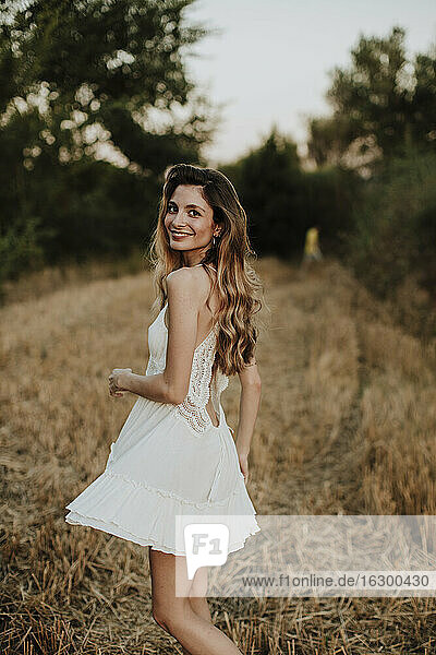 Lächelnde schöne Frau in weißem Kleid steht im Weizenfeld bei Sonnenuntergang