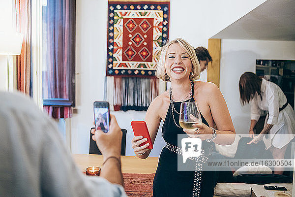 Mann fotografiert fröhliche Freundin  die ihr Smartphone hält  während sie auf einer Party Wein trinkt