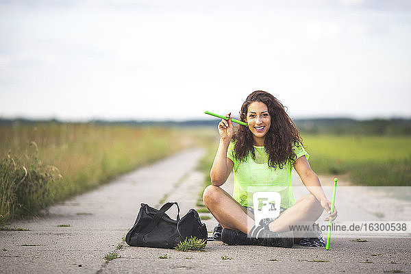 Young woman holding drumsticks while sitting on road against sky