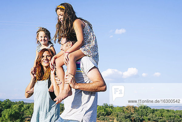 Glückliche Eltern tragen ihre Töchter auf den Schultern gegen den Himmel an einem sonnigen Tag