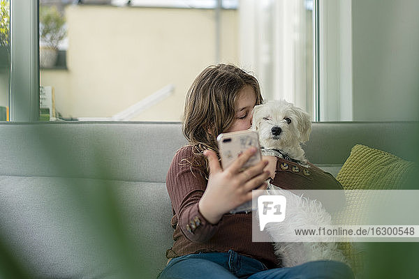 Mädchen  das ein Selfie mit dem Smartphone macht  während es den Hund auf dem Sofa küsst
