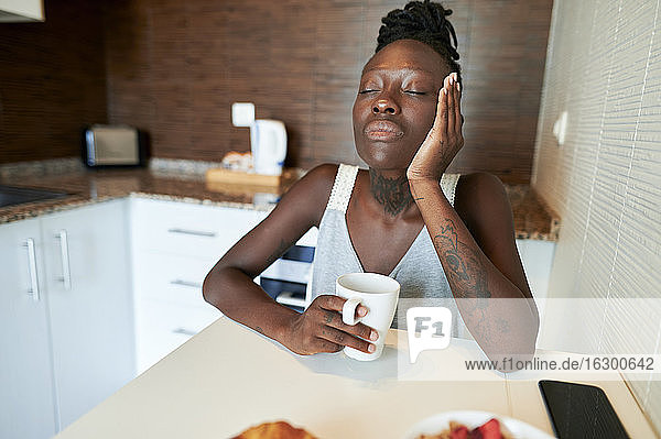 Junge Frau mit geschlossenen Augen sitzt mit Kaffeetasse am Tisch in der Küche