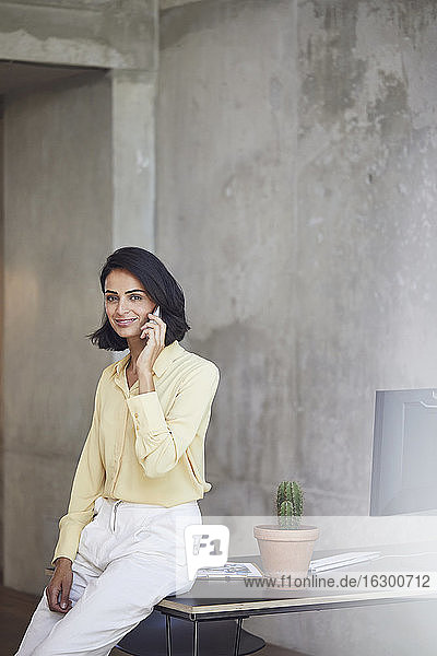 Smiling businesswoman talking over mobile phone while sitting on desk against wall in office