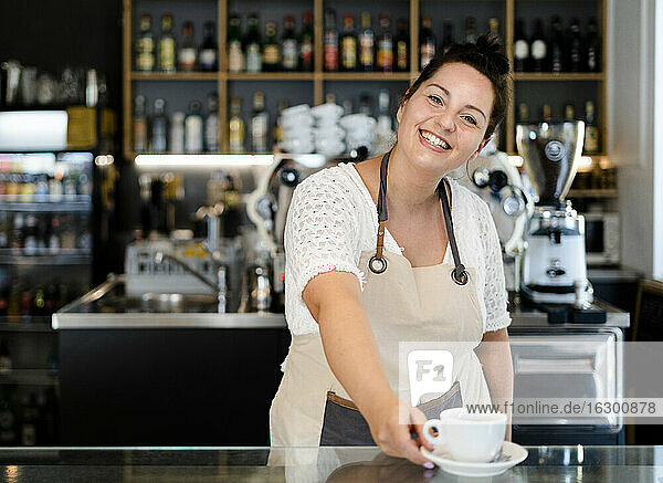 Lächelnde Besitzerin  die eine Kaffeetasse auf dem Tresen serviert  während sie in einem Café arbeitet