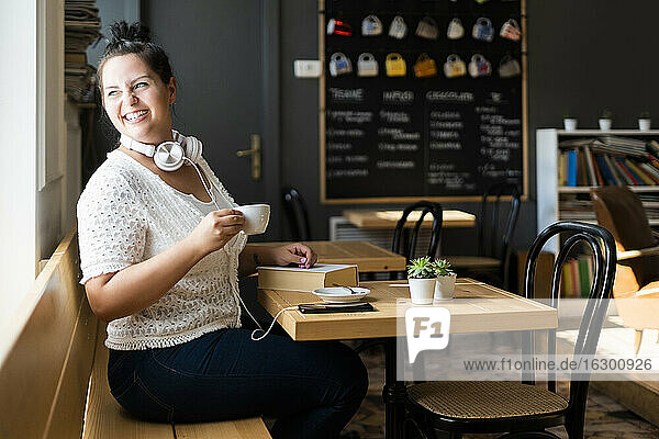 Smiling woman holding coffee cup looking through window while sitting at table in cafe