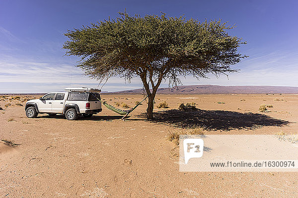 Mann ruht sich in einer Hängematte aus  die zwischen einem einsamen Wüstenbaum und einem Geländewagen hängt