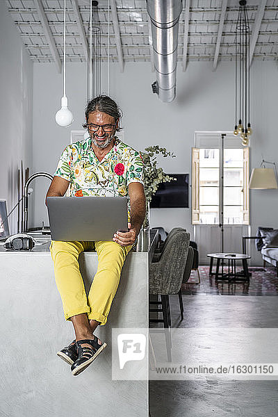 Smiling man using laptop while sitting on counter at home