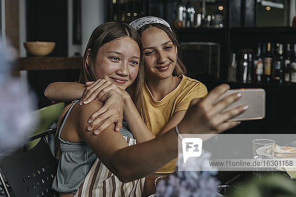 Teenage girlfriends sitting at dining table  embracing and taking smartphone selfies