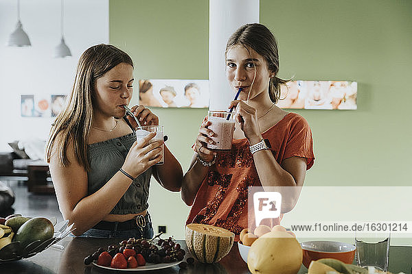 Teenage girls standing in kitchen drinking fresh fruit smoothies with drinking straws
