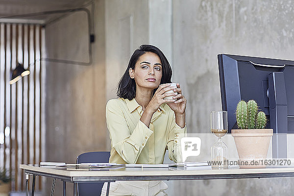 Nachdenkliche Geschäftsfrau  die eine Kaffeetasse hält  während sie am Schreibtisch im Büro sitzt