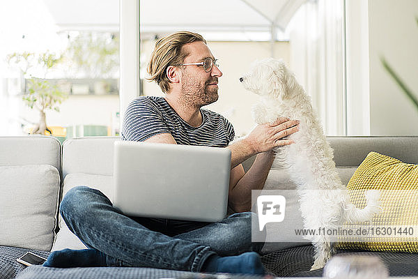 Mann mit Laptop  der einen Hund hält  während er auf dem Sofa sitzt