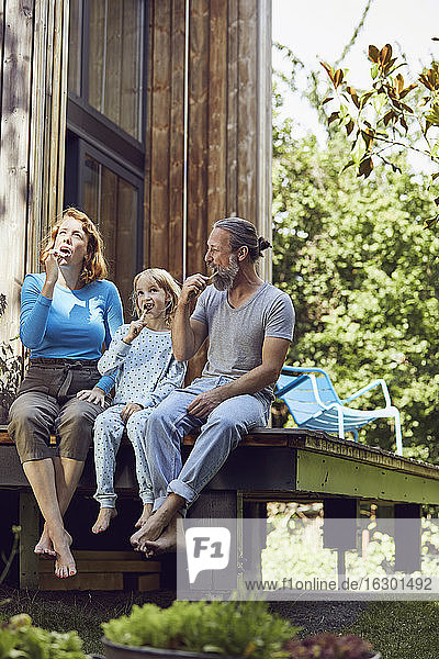 Tochter mit Eltern beim Zähneputzen  während sie vor einem kleinen Haus sitzen