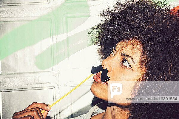 Close-up of woman with curly hair holding fake mustache on face against wall