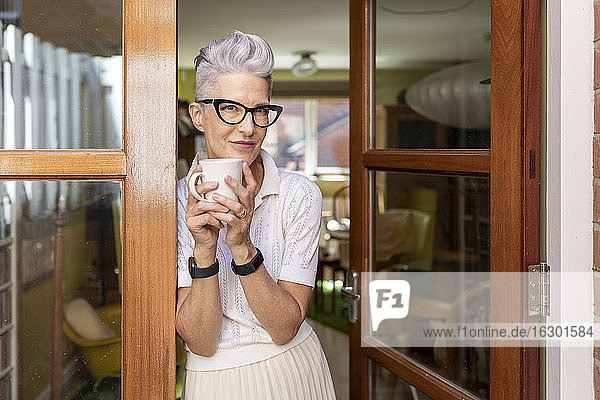 Ältere Frau lehnt an der Tür und hält eine Kaffeetasse in der Hand