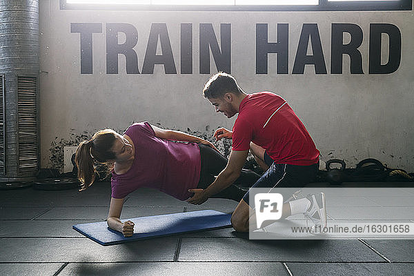 Fitnesslehrer  der einer Sportlerin beim Üben der Plank-Position auf einer Matte im Fitnessstudio hilft