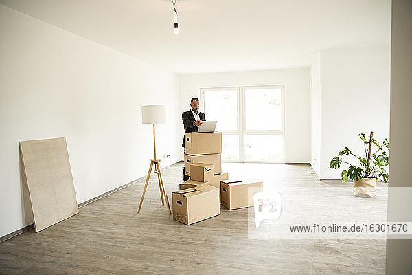 Businessman working over laptop on stacked boxes while standing in new apartment
