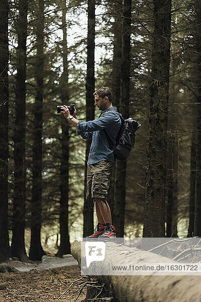 Male hiker photographing with camera while standing on log in woodland