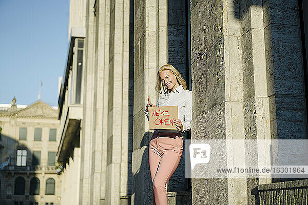 Lächelnde weibliche Fachkraft  die ein offenes Schild aus Pappe an einem Gebäude im Finanzbezirk hält