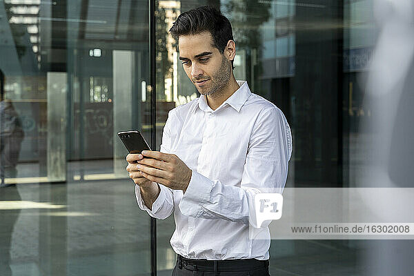 Male professional using smart phone while standing against window in office