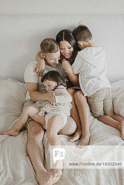 Smiling parents embracing kids while sitting on bed in bedroom