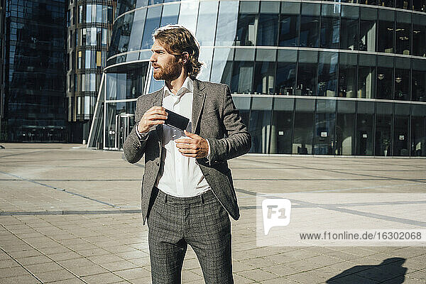 Young businessman looking away while holding smart phone against office building in downtown