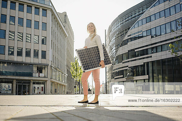 Lächelnde schöne Unternehmerin  die ein Solarmodul hält  während sie auf einem Fußweg im Finanzviertel steht