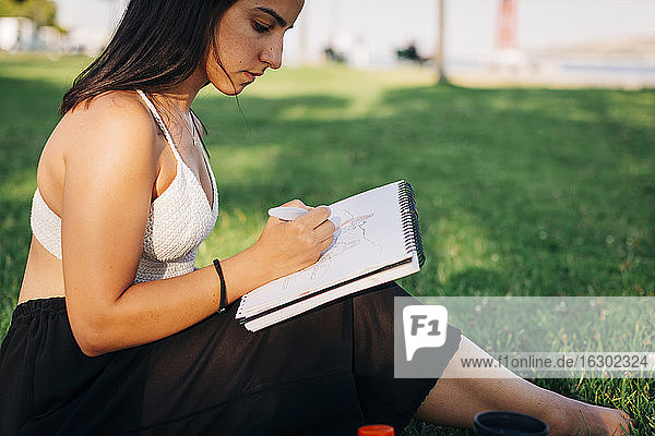Schöne Frau malt auf einem Buch  während sie in einem öffentlichen Park sitzt