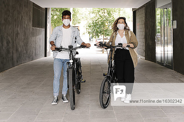 Couple in protective face masks standing with electric bicycles on footpath