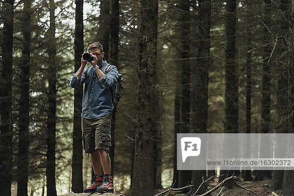 Male hiker photographing with camera while standing against trees in forest