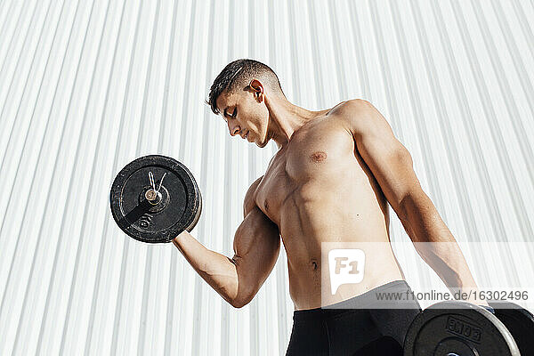Shirtless athlete lifting dumbbell while standing against wall