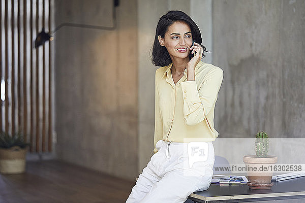 Lächelnde Unternehmerin  die über ein Mobiltelefon spricht  während sie auf einem Schreibtisch im Büro sitzt
