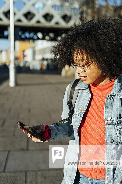 Nahaufnahme einer Frau mit lockigem Haar  die ein Mobiltelefon benutzt  während sie an einem sonnigen Tag auf dem Gehweg steht
