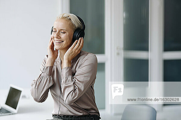 Lächelnde Geschäftsfrau hört Musik über Kopfhörer  während sie im Büro sitzt