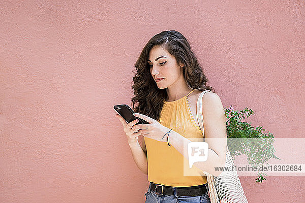 Schöne junge Frau  die ein Smartphone benutzt  während sie mit einer wiederverwendbaren Netztasche vor einer rosa Wand steht