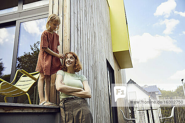 Mädchen bindet ihrer Mutter die Haare  während sie vor einem kleinen Haus steht