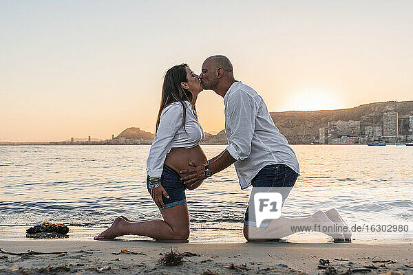 Älterer Mann küsst schwangere Frau  während er bei Sonnenuntergang am Strand kniet