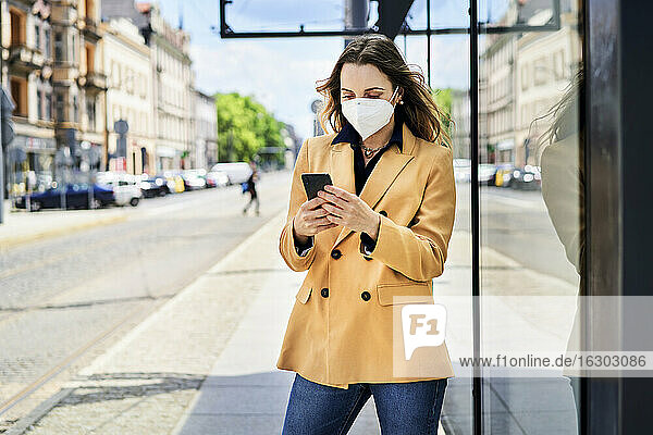 Frau mit Schutzmaske benutzt ihr Smartphone beim Warten an der Straßenbahnhaltestelle
