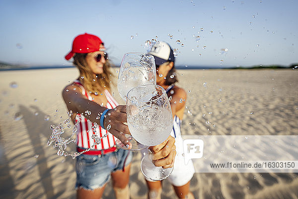 Mid adult female friends splashing drink while toasting at beach on sunny day
