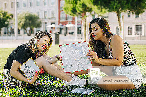 Young woman showing map to female friend while sitting on grassy land in park