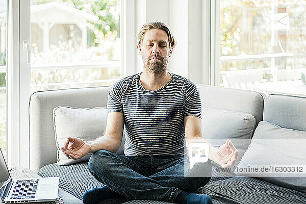Male freelancer meditating by laptop on sofa