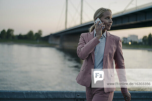 Female professional looking away while talking on smart phone against bridge at dusk