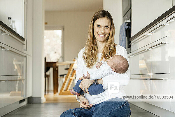 Blond woman holding sleeping baby boy while sitting on floor at home