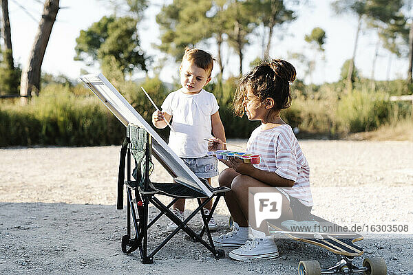 Baby Mädchen malen mit Schwester auf Leinwand im Park bei Sonnenuntergang
