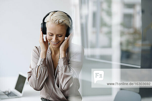 Lächelnde Unternehmerin mit geschlossenen Augen  die über Kopfhörer Musik hört  während sie in einem Loft sitzt