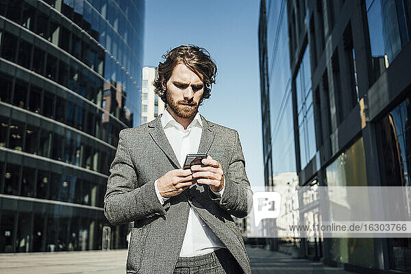 Männlicher Fachmann  der ein Mobiltelefon benutzt  während er an einem modernen Bürogebäude in der Innenstadt an einem sonnigen Tag steht
